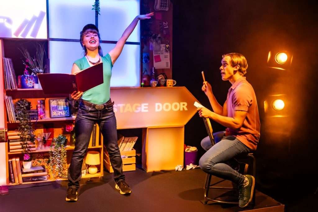 Heidi sings with a score in one hand and an arm raised. Jeff conducts from a nearby chair.