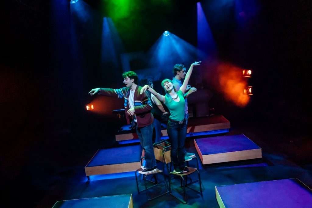The four friends stand on chairs waving their arms in a floating motion as if flying