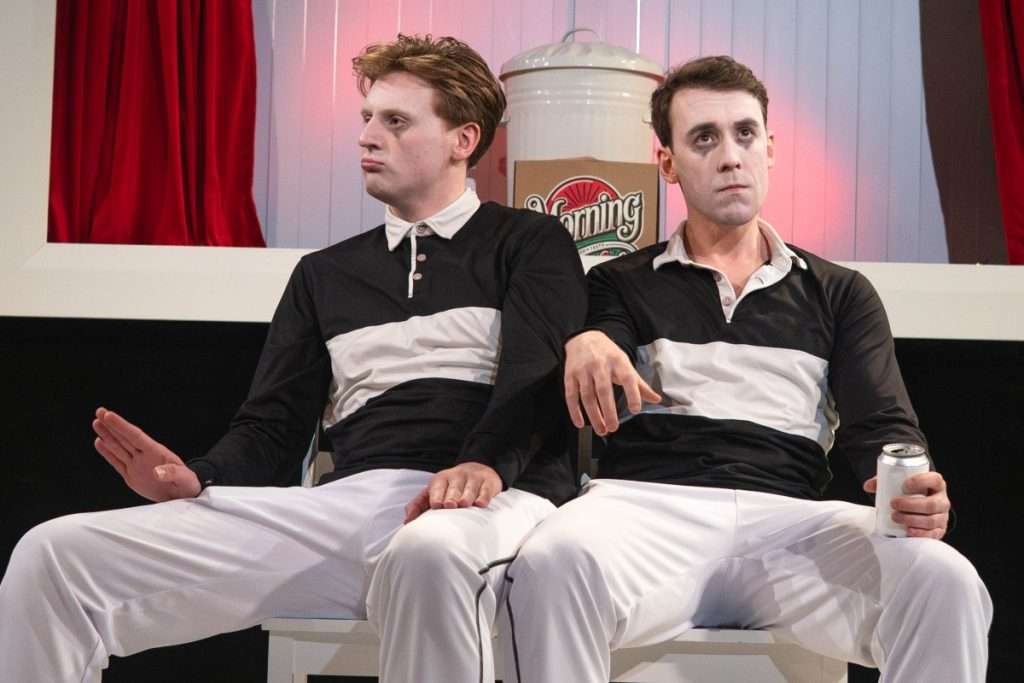 Nick Hyde and Oliver Maynard wear matching outfits and clown make up, they sit looking bored