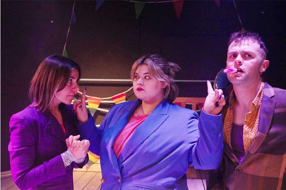 Mali Wen Davies wears a blue blazer and holds her fingers up to the lips of Jude St James and James Lowrie
