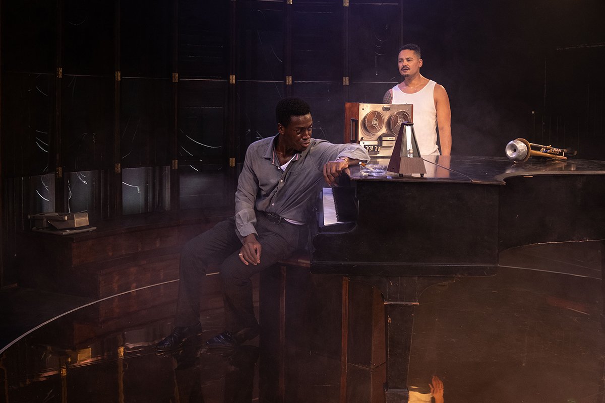 Miles (Benjamin Akintuyosi) and Jay (Jay Phelps) by a large grand piano. Haze blurs the image.