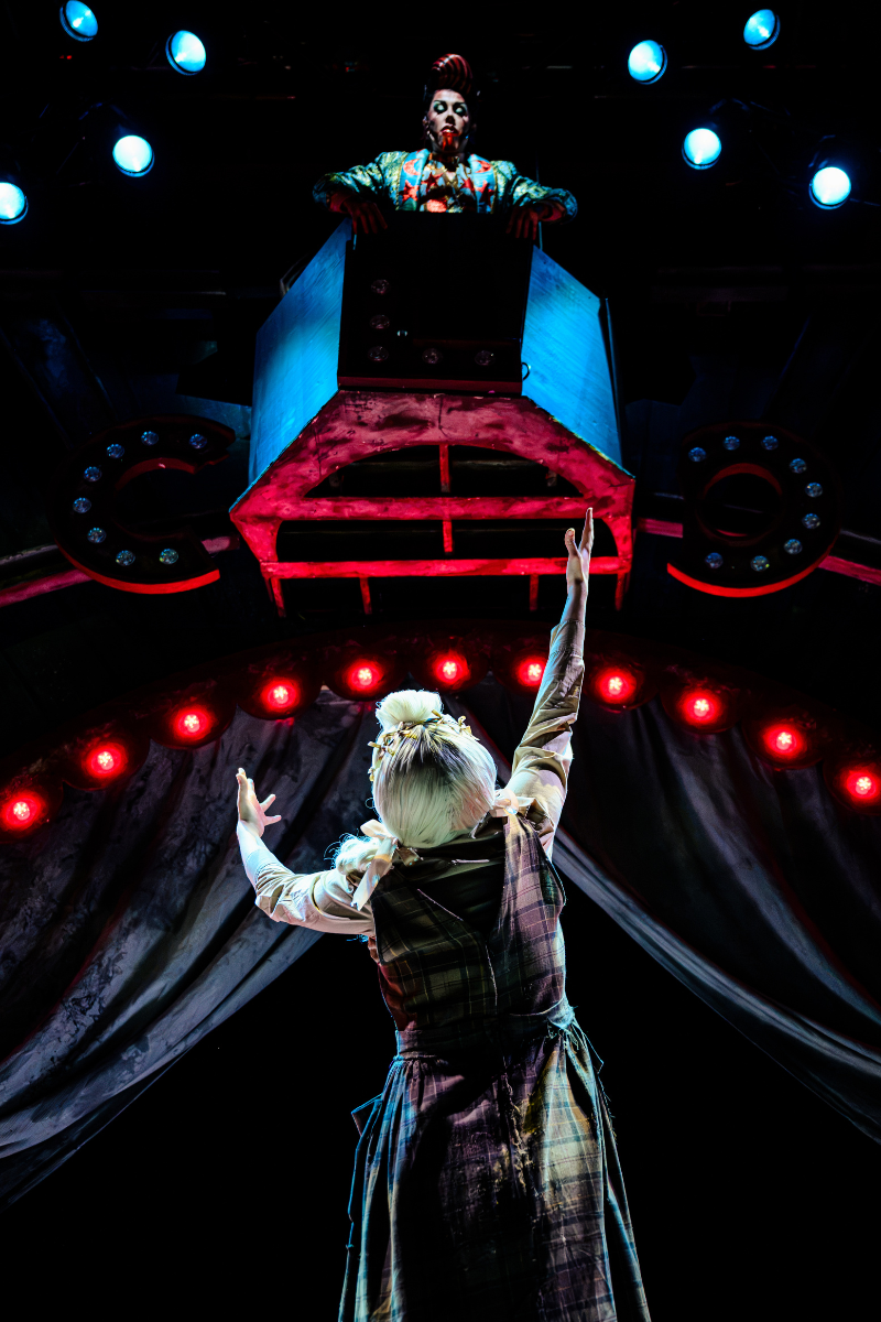 Grace Galloway as Jane Doe, photographed from behind, reaches her arms up to Karnak (played by Divina de Campo) who is situated in a balcony box above.